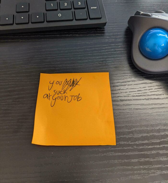 Orange sticky note with handwritten message on a dark desk near keyboard and blue trackball, representing funny work-from-home memes.