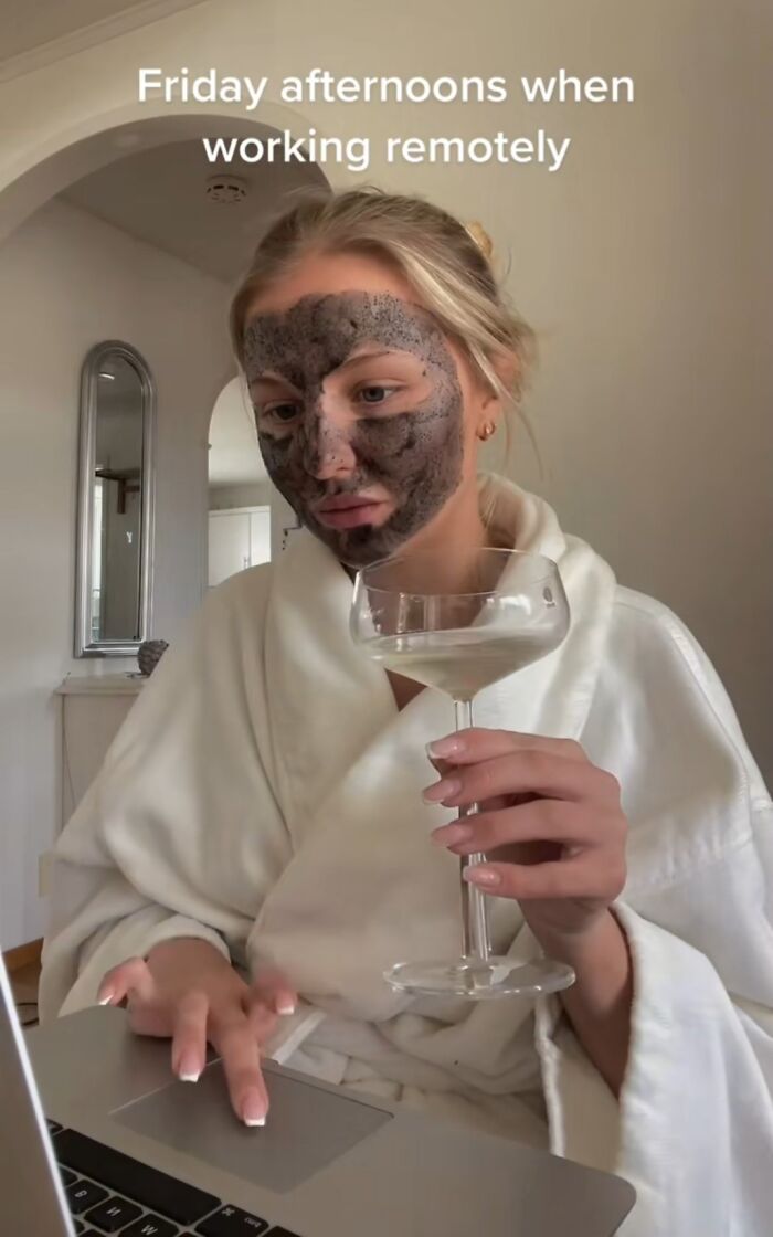 Young woman with a face mask and bathrobe working on laptop while holding a wine glass, illustrating funny work-from-home memes.