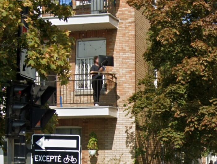 Person working from home on a balcony holding a computer monitor, surrounded by trees and urban buildings.