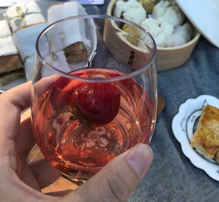 Hand holding a glass of pink drink with a strawberry, with picnic foods in the background for International Picnic Day.