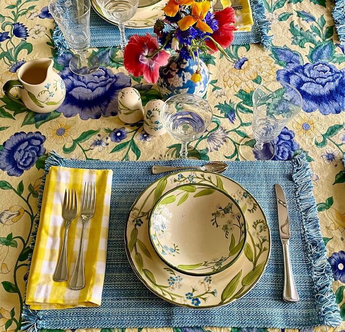 Table setting with floral plates, cutlery, checkered napkin, and glassware on a colorful floral tablecloth for an al fresco feast.