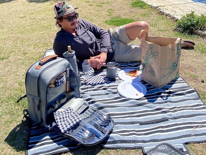 Man relaxing on grass with picnic essentials including cooler bag, utensils, drinkware, and food for an al fresco feast.