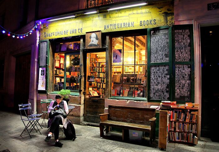 Cozy antiquarian bookstore at night with warm lighting, wooden benches, and books displayed outside among stunning bookstores.