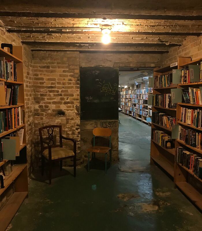 Cozy underground bookstore with exposed brick walls, wooden shelves filled with books, and vintage chairs in warm lighting.