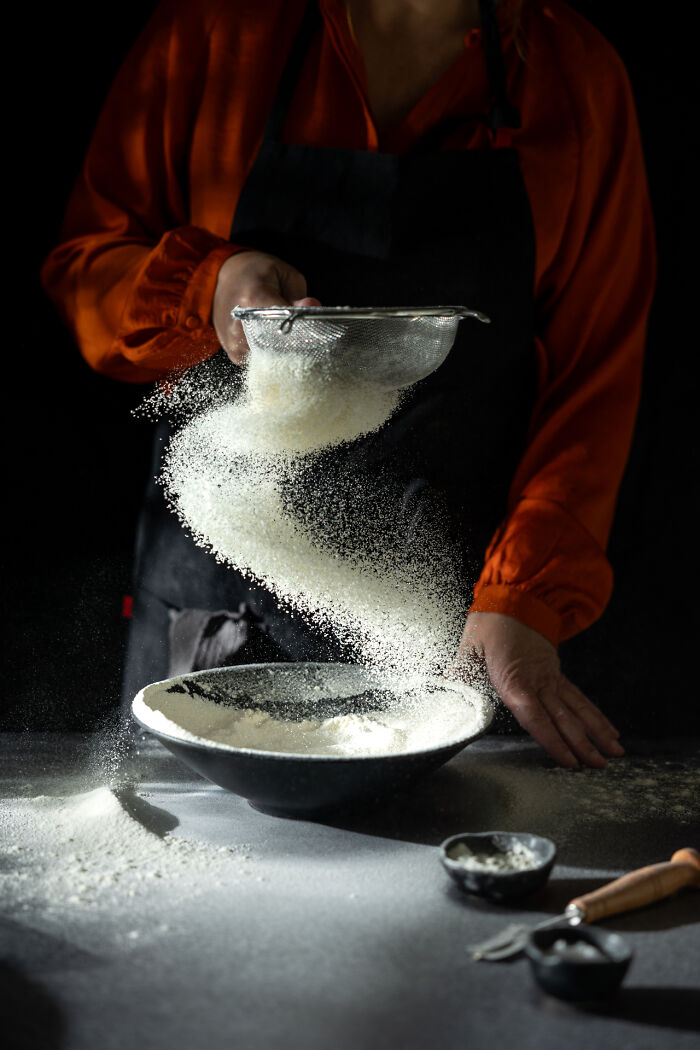 Person sifting flour into bowl with motion blur, showcasing creative food photography techniques for World Food Photography Awards.