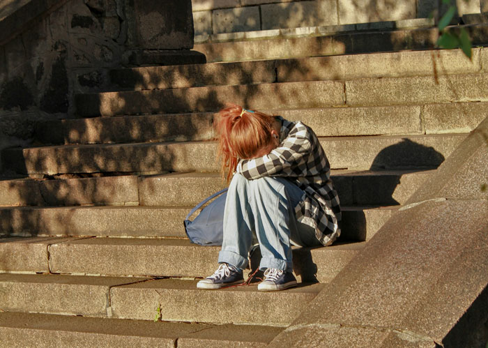 Teen sitting on stone steps with head down, wearing casual clothes, depicting stress from bosses, teachers, or parents.