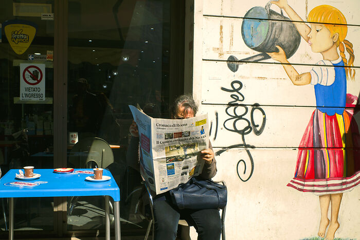 Street photography moment showing a person reading a newspaper beside a colorful mural of a girl watering plants.