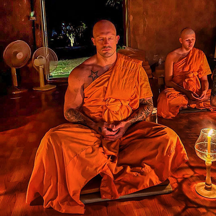 Former pole dancer wearing orange monk robes, meditating in a dimly lit room with another monk in the background.