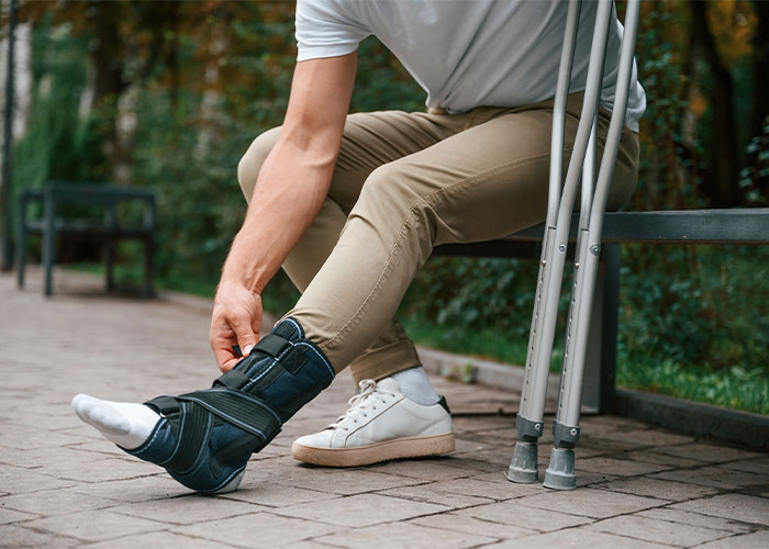 Person with leg injury wearing an ankle brace and adjusting it while resting on a bench with crutches nearby.