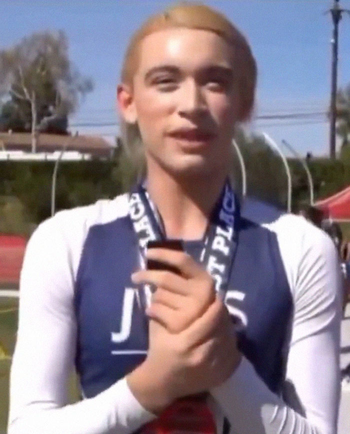 High school athlete wearing first-place medal, standing outdoors at track event with a focused expression.