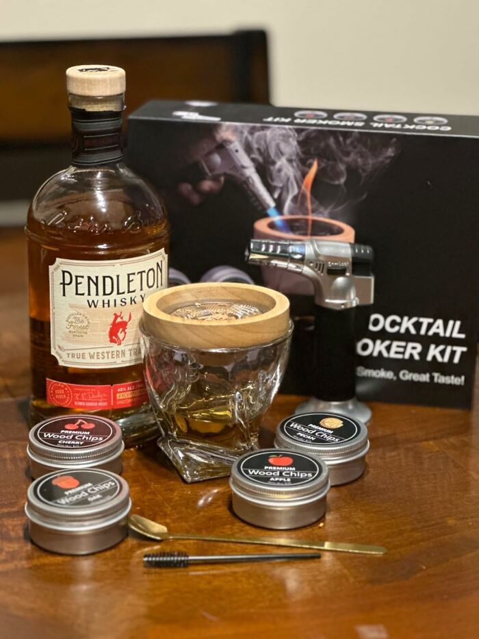 Father's Day gifts including Pendleton whiskey, cocktail smoker kit, wood chips, and a skull-shaped glass on a wooden table.