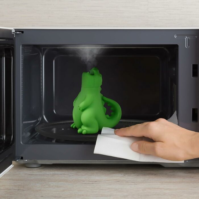 Green dinosaur-shaped microwave steam cleaner emitting steam while a hand wipes the microwave interior, showcasing unique purchases.