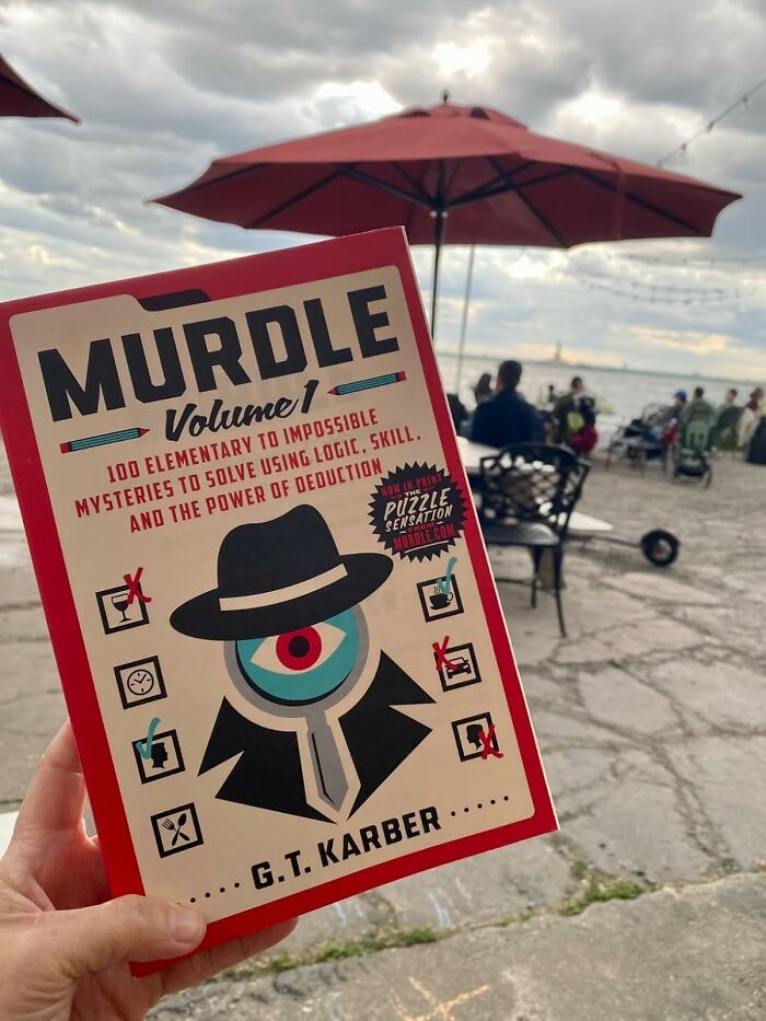 Hand holding Murdle Volume 1 puzzle book outdoors near a waterfront café, ideal Father's Day finds for dads who love puzzles.
