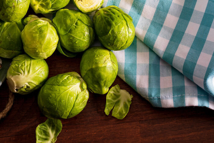 Fresh Brussels sprouts with water droplets on a wooden surface next to a blue and white checkered kitchen towel.