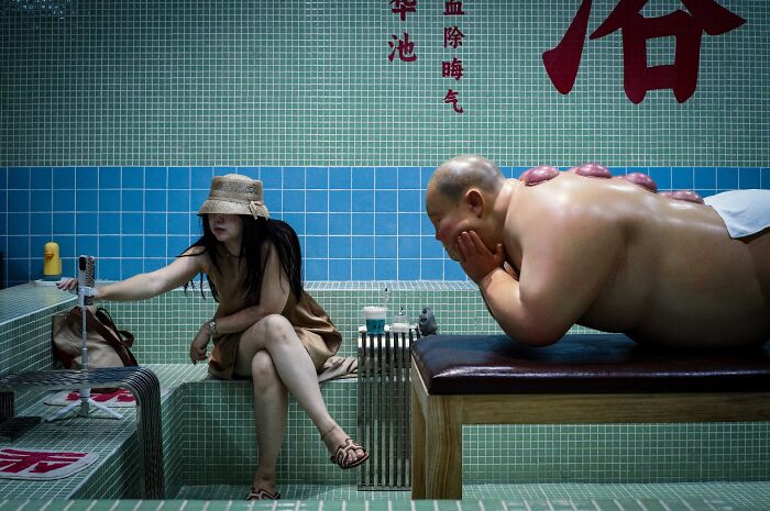 Woman in a hat taking a photo with a phone while a man receives a massage in a tiled room, street photography grant scene.
