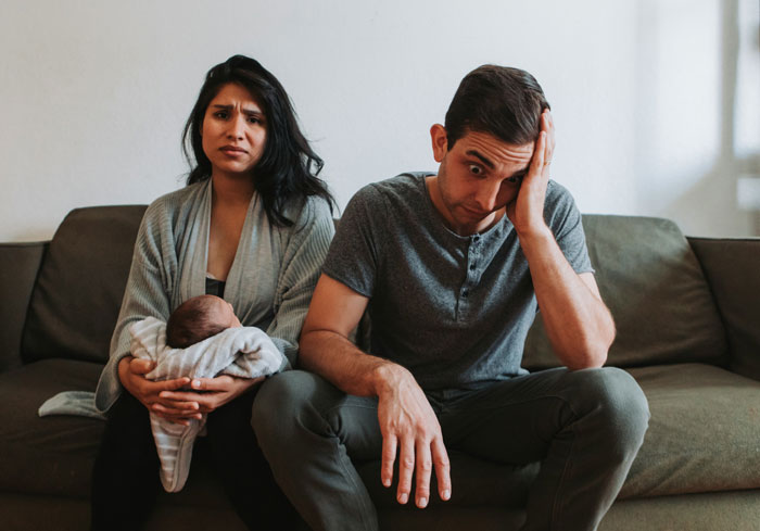 Woman holding baby looking concerned beside stressed boyfriend on couch, reflecting conflicting feelings about having kids and marriage Woman holding baby looking concerned beside stressed boyfriend on couch, reflecting conflicting feelings about having kids and marriage