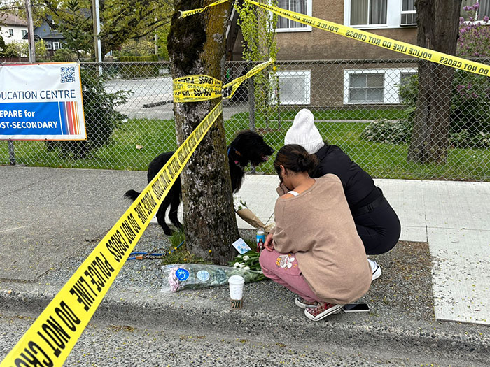 Two people mourn at a Vancouver attack scene with police tape, placing flowers near a tree on the sidewalk.