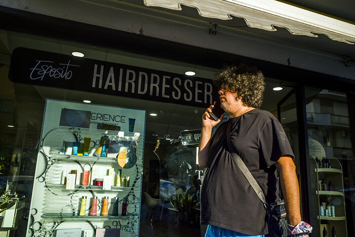 Man with curly hair standing outside a hairdresser shop, captured in a vibrant street photography moment.