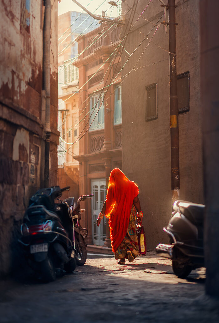 Woman in vibrant orange sari walking down narrow street with scooters, a striking street photography moment captured.