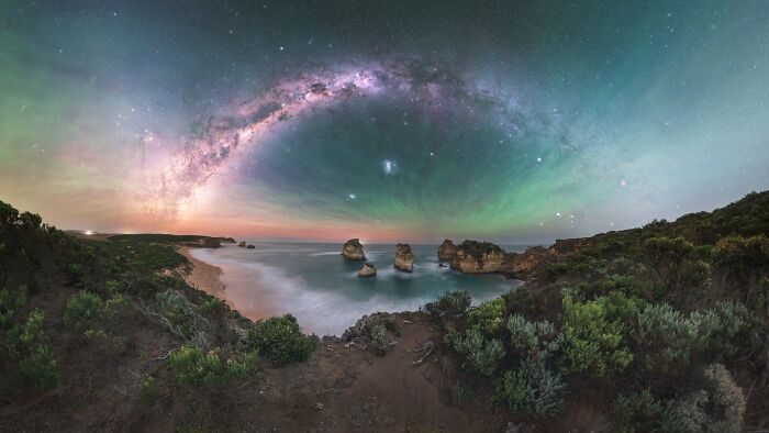 Panoramic night sky image featuring a vibrant Milky Way arch over a coastal landscape with rock formations.