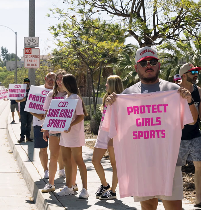 Protesters holding signs supporting girls sports stand outdoors in a sunny area advocating protection for female athletes.