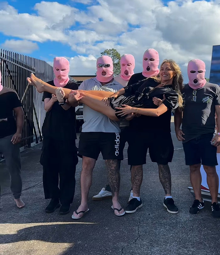 Adult content creator smiling while being held by a group wearing pink masks, linked to bleeding hospital update story.
