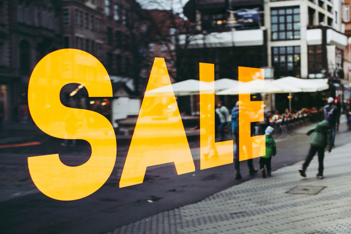 Yellow sale sign on a glass window reflecting people and a city street, illustrating common money loss mistakes.