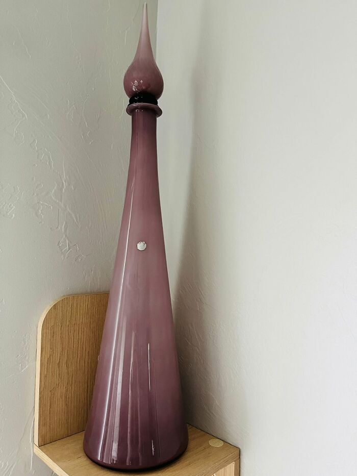 Tall purple vintage decanter with a pointed stopper displayed on a wooden shelf among thrifted finds.