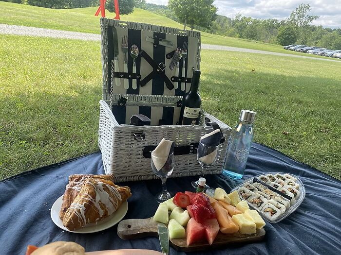 Picnic basket with utensils, wine, fruit platter, sushi, and croissants set on a blanket for an al fresco feast outdoors.