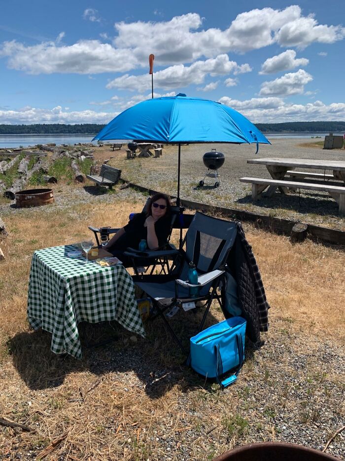 Woman relaxing under a blue umbrella at a picnic setup with a green checkered tablecloth near a waterfront on a sunny day.