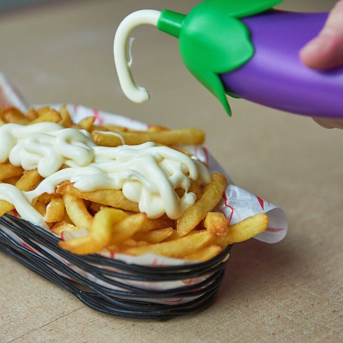 Funny kitchen gadget dispensing mayonnaise over crispy fries in a black basket lined with paper on a beige surface.