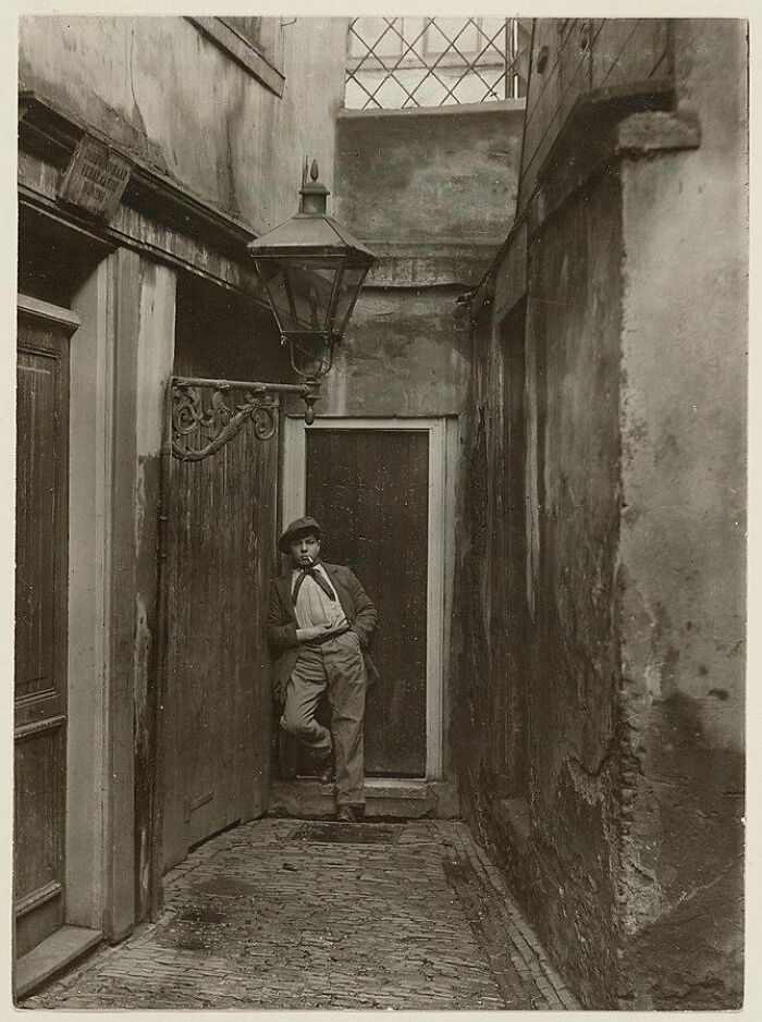 Young boy smoking a pipe, leaning against a door in a narrow historic alley showing unseen side of history.
