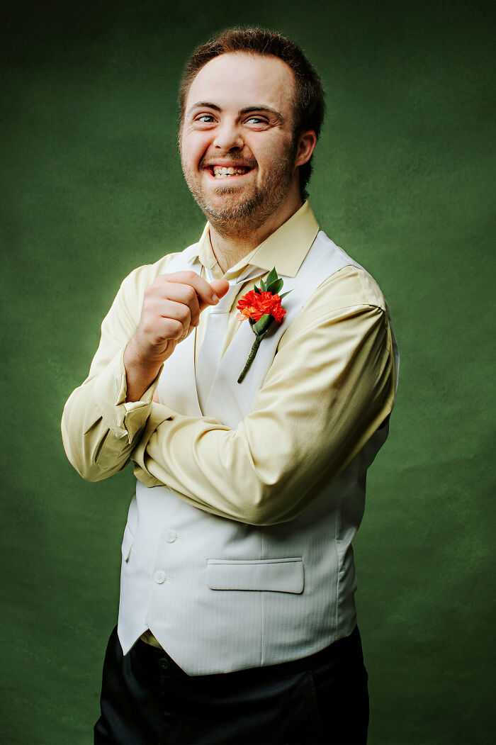 Young man with Down syndrome smiling confidently, wearing a dress shirt and vest with a red flower boutonniere.