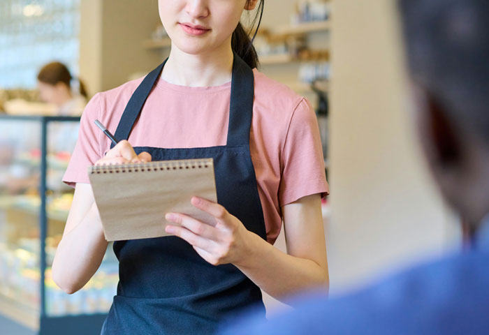 Young woman in a pink shirt and black apron taking notes, illustrating problematic behaviors women get a pass for according to men.