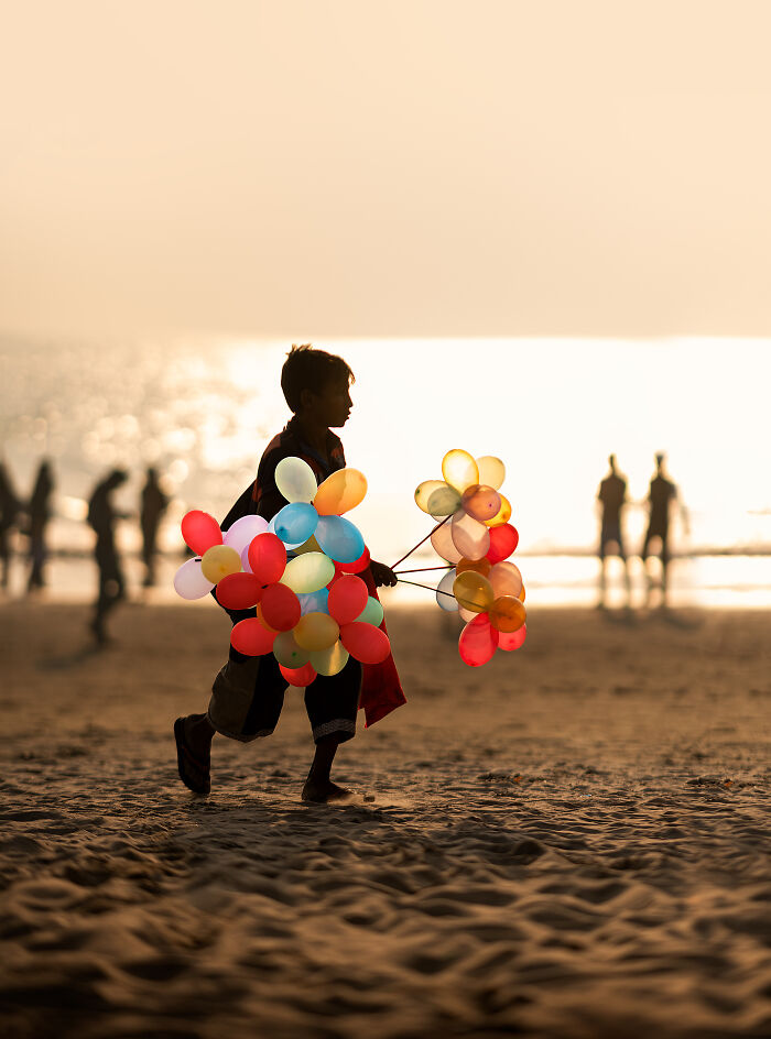 Silhouette of a child holding colorful balloons walking on the beach at sunset in a street photography moment.