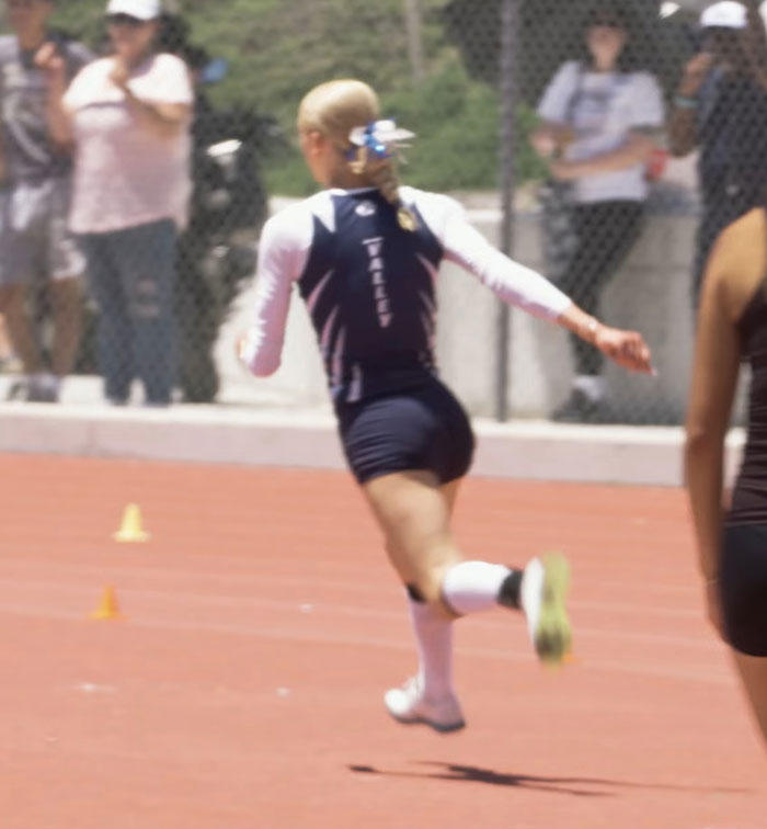 High school girl athlete sprinting on track during race with spectators behind a chain-link fence on a sunny day