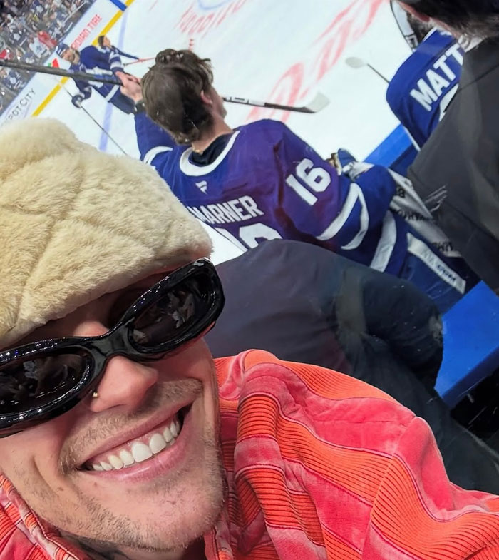 Justin Bieber smiling at a hockey game, wearing sunglasses and a furry hat, with players on the ice in the background. Justin Bieber smiling at a hockey game, wearing sunglasses and a furry hat, with players on the ice in the background.