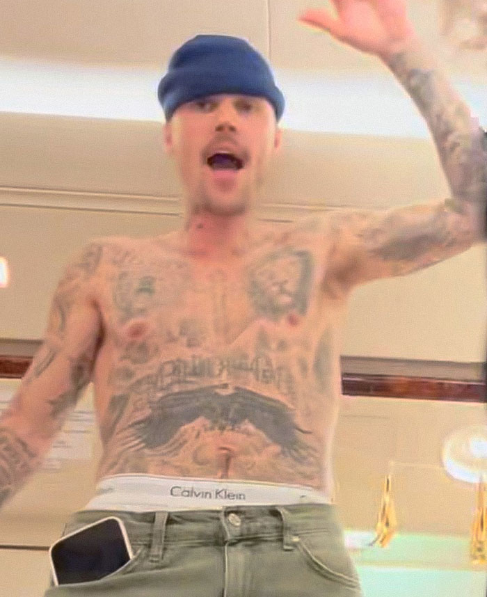 Justin Bieber shirtless in LA, showing tattoos and wearing a beanie while expressing emotion during casual moment indoors.