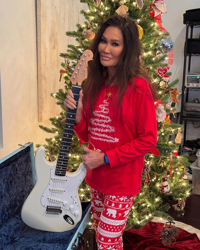 Tia Carrere, 90s icon, posing with guitar in festive red holiday outfit near decorated Christmas tree.