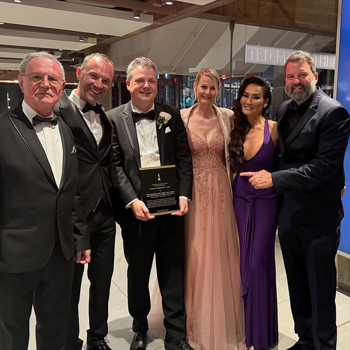 Tia Carrere in a plunging purple gown posing with five people at an event, showcasing her timeless '90s icon style.