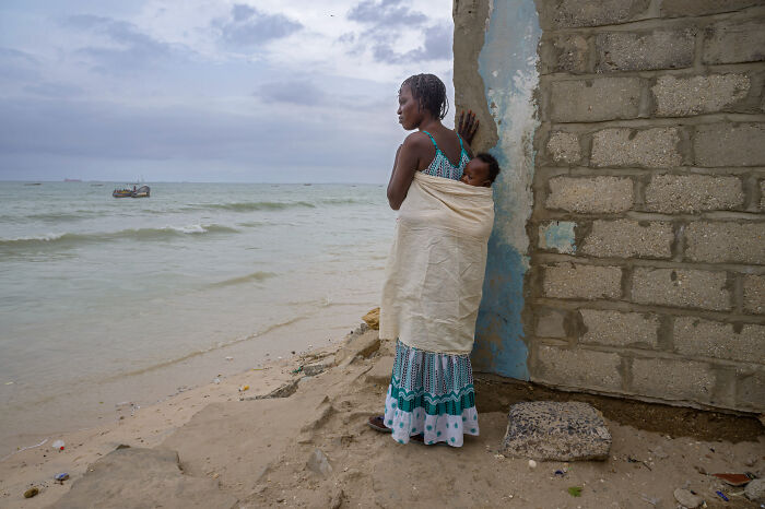 Woman carrying baby on her back, standing by a weathered wall and looking out over the ocean, capturing essence of humanity.