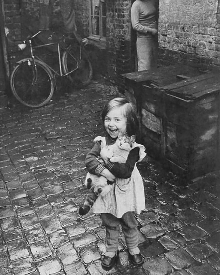 Vintage photo of a joyful child holding a cat, capturing the timeless bond between kids and cats on cobblestone street.