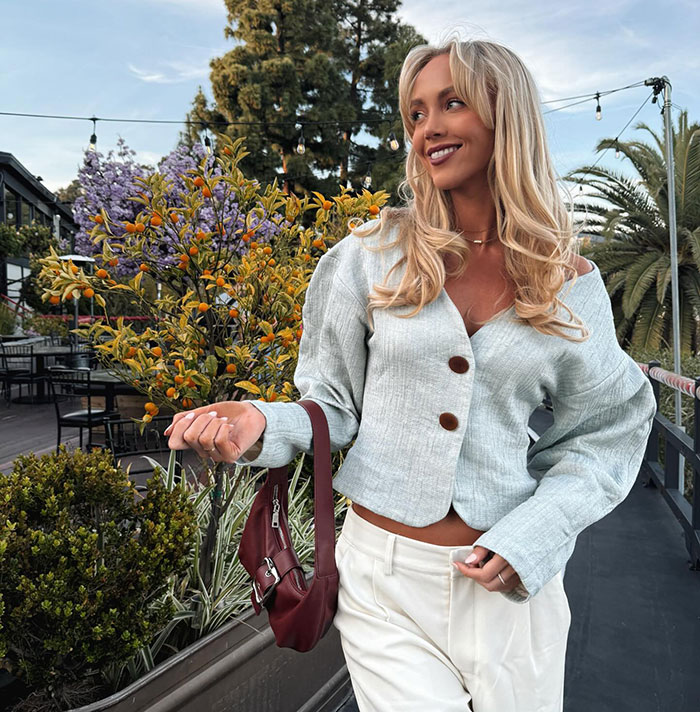 Adult star Annie Knight smiling outdoors, wearing a light jacket and holding a maroon purse near colorful plants. Adult star Annie Knight smiling outdoors, wearing a light jacket and holding a maroon purse near colorful plants.
