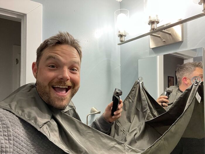 Man smiling while holding a beard trimming apron and clipper, demonstrating a Shark Tank pitch product at home.