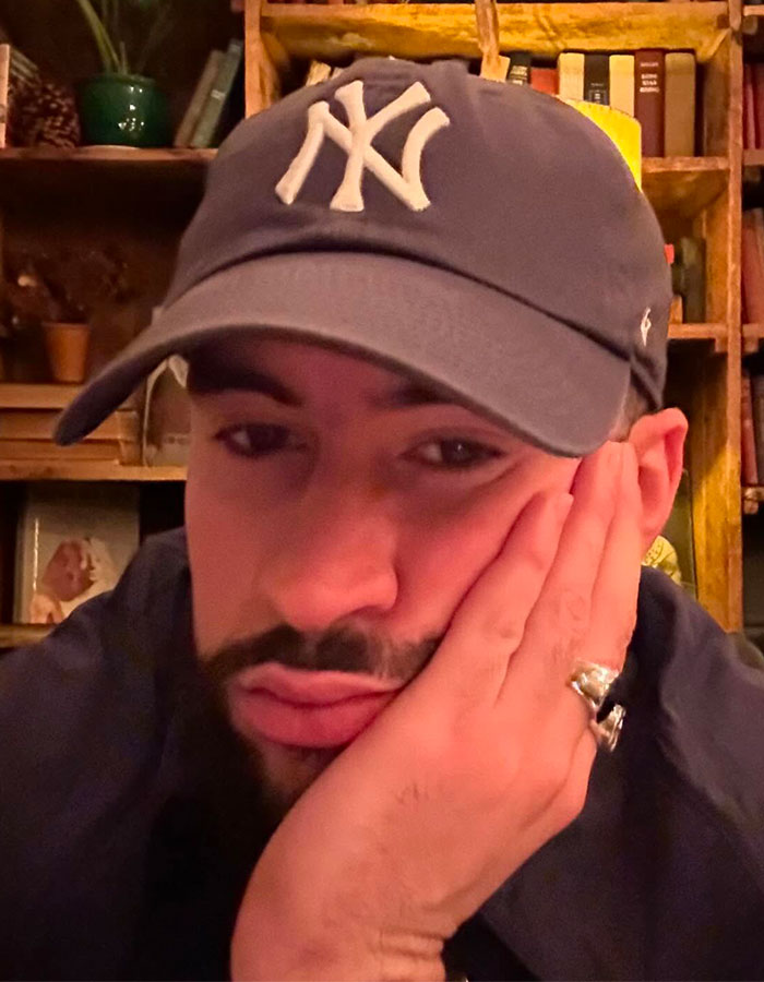 Close-up selfie of Bad Bunny wearing a New York Yankees cap, resting his face on his hand indoors with bookshelves behind.