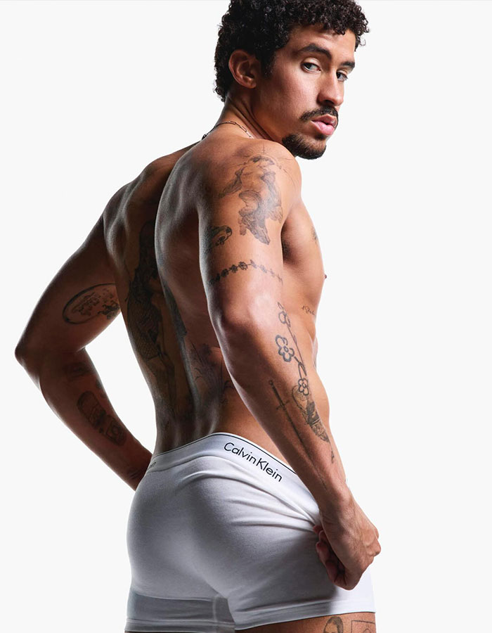 Male model with tattoos wearing revealing white Calvin Klein underwear posing against a white background.