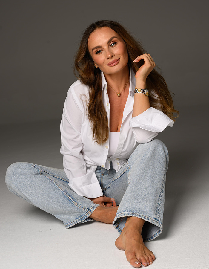 Female cosmetologist with long hair in white shirt and jeans sitting barefoot, representing cosmetologist insights on Hollywood stars.