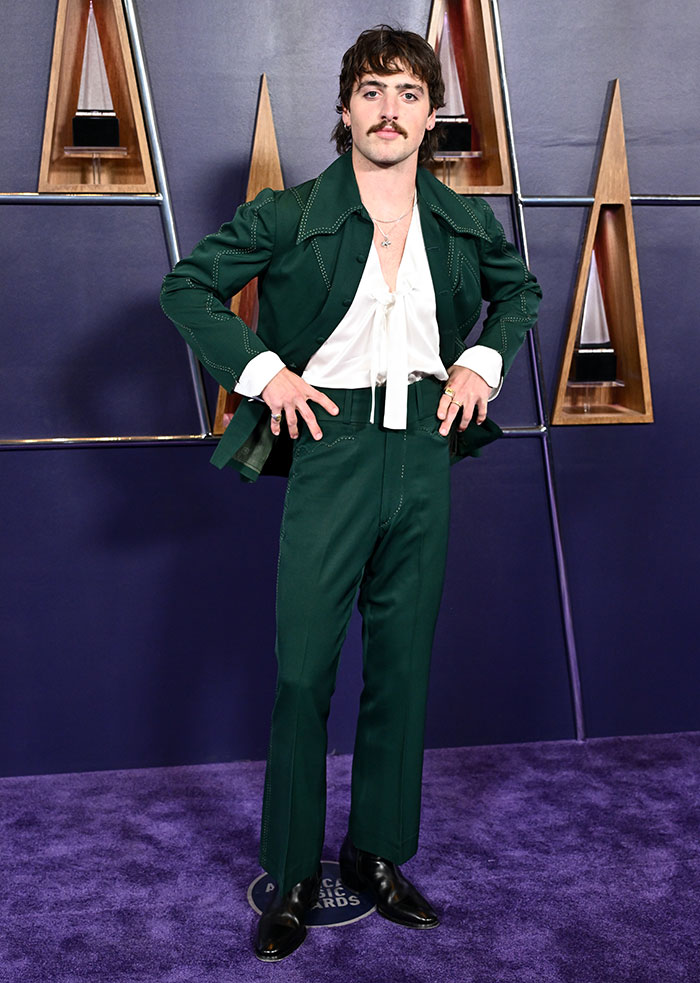 Man posing on purple carpet wearing green suit and white blouse, representing worst dressed stars from 2025 American Music Awards.