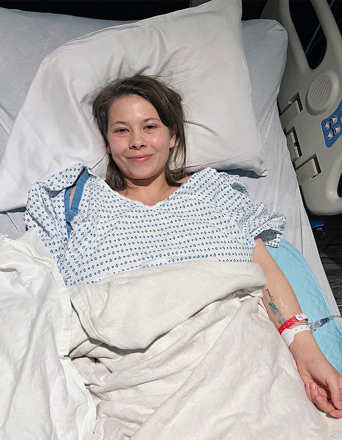 Bindi Irwin in hospital bed wearing a gown, giving a health update after a medical emergency from incurable disease.