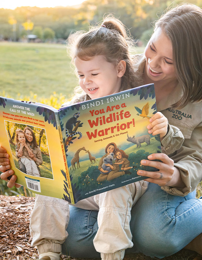 Bindi Irwin reading a wildlife book with a child outdoors, sharing a joyful moment about animals and nature conservation.